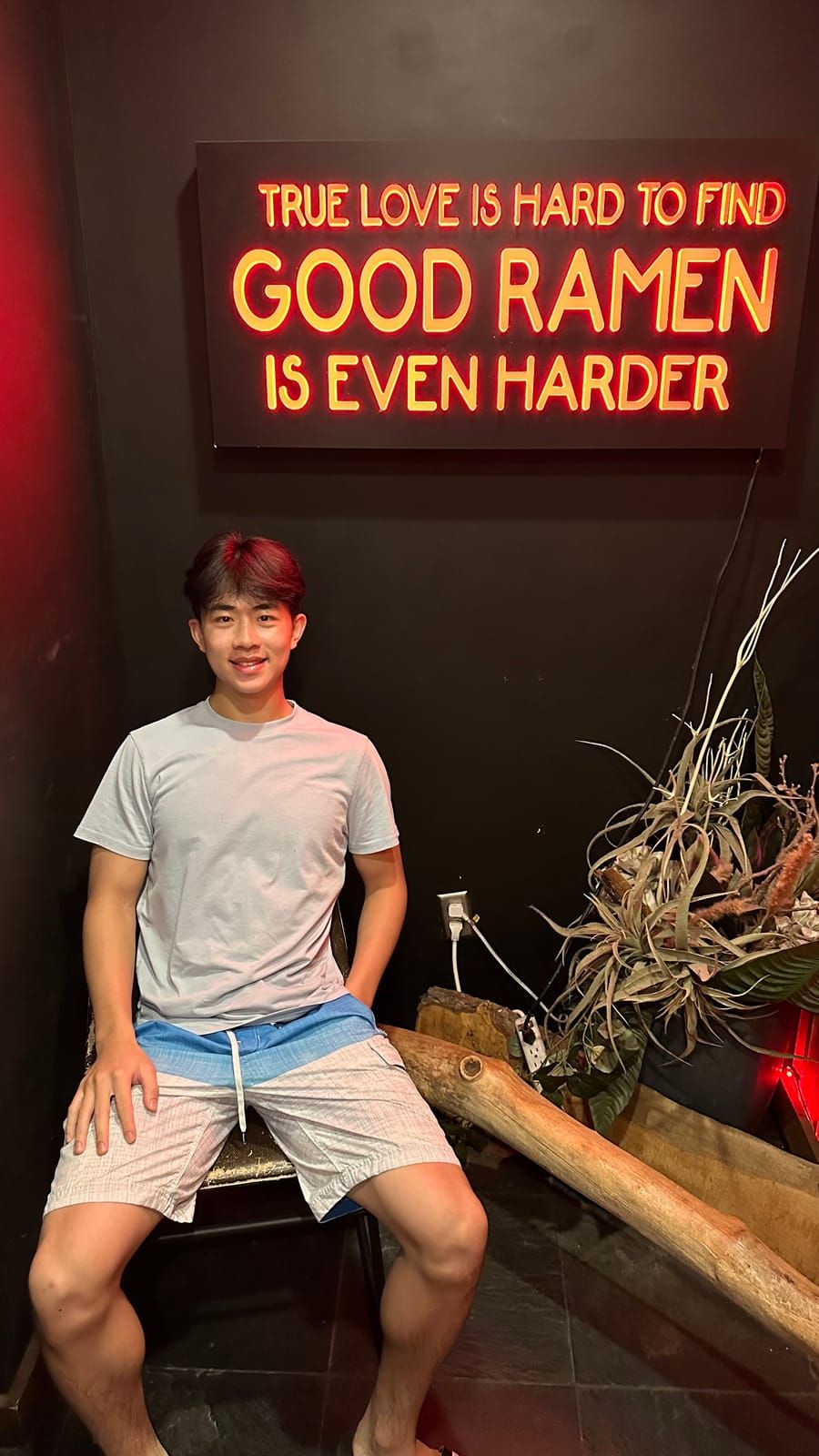 Joshua sitting beneath a neon sign that reads “good ramen is even harder”